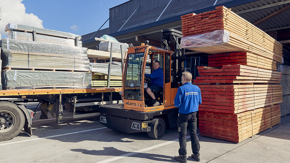 Un homme conduit un chariot élévateur orange, transportant une pile de planches de bois. Un autre homme, en tenue de travail bleue, se tient à côté, supervisant ou attendant. À gauche, un camion est chargé de matériaux emballés, probablement prêts pour le